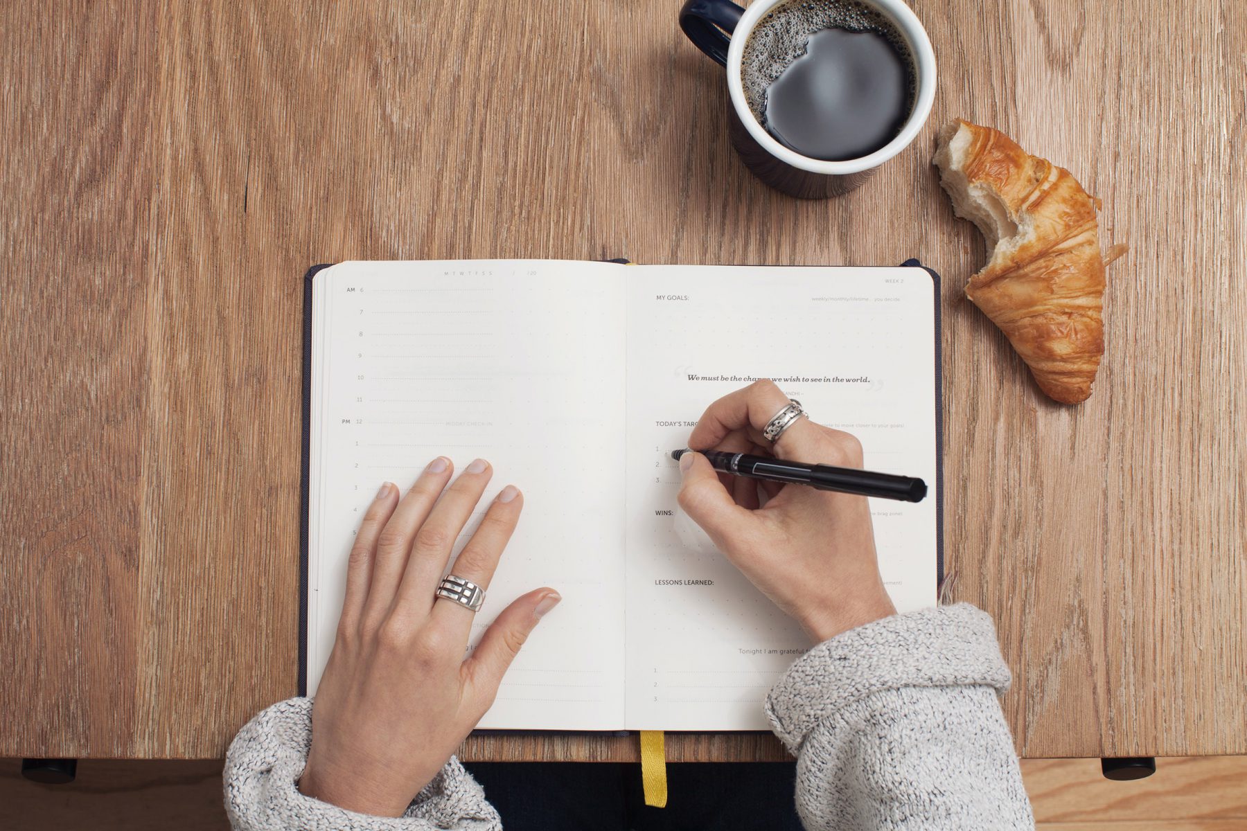 Hands writing in a journal on a wooden table with a cup of coffee and croissant 