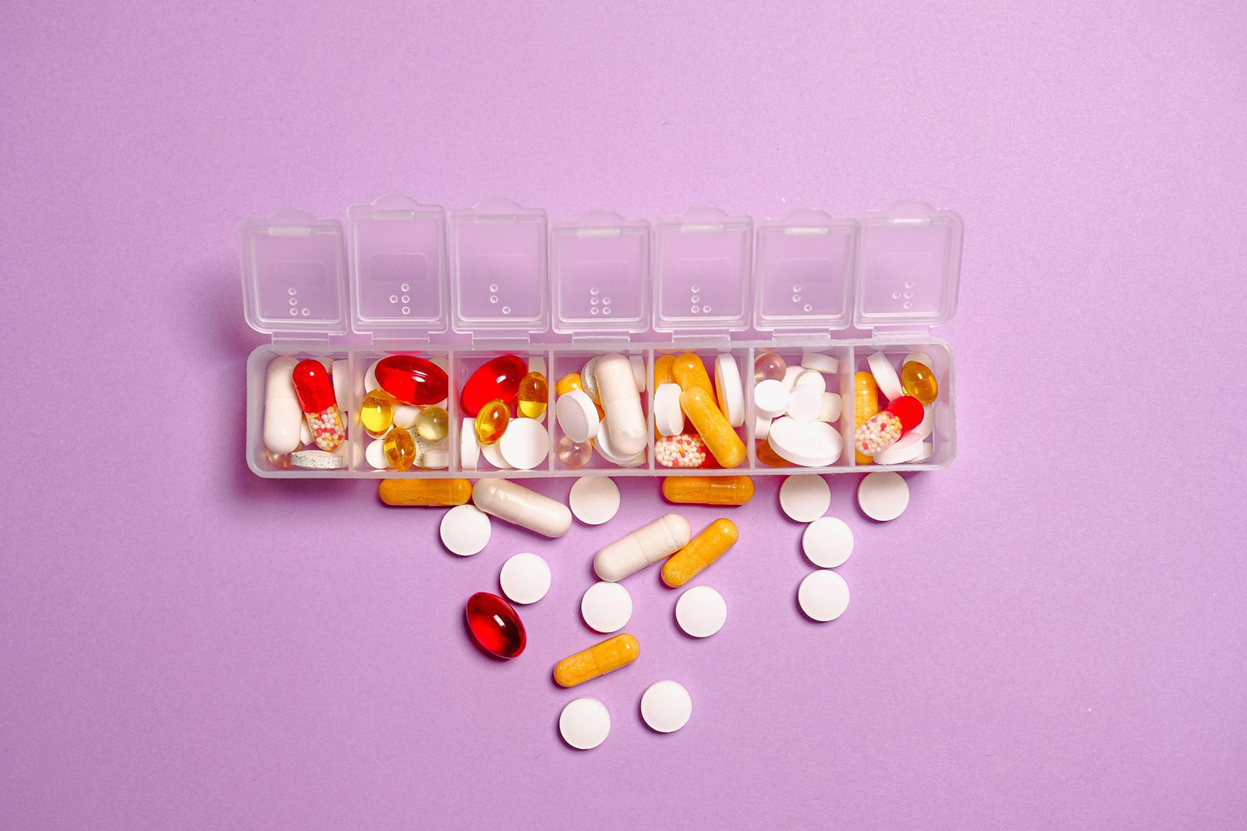 A weekly pill container overflowing with vitamins on a purple background
