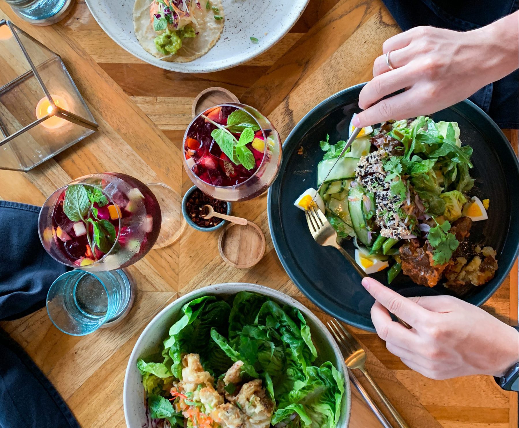 bowls of salad, two hands with cutlery and two fruity drinks on wooden table